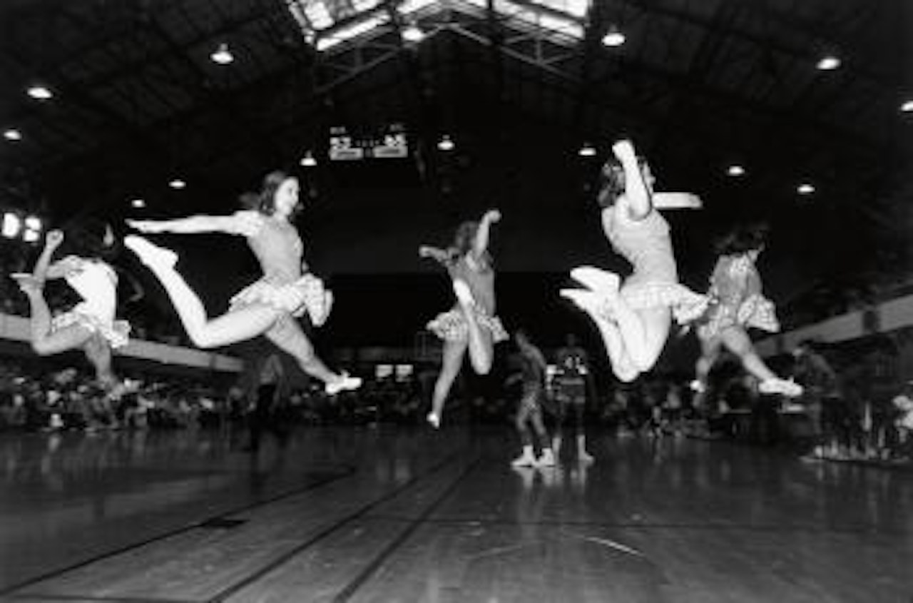 A cheerleading squad in formation, mid-flight, gingham skirts flouncing as the basketball team behind them make their way to the bench by Garry Winogrand